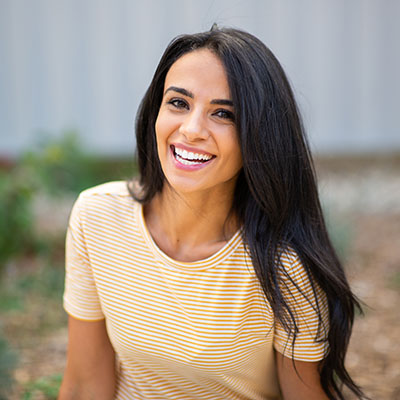 A woman with long dark hair smiling at the camera, wearing a yellow top and sitting on the ground.
