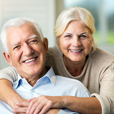 The image shows an older man and woman smiling and embracing each other, with the man wearing glasses and a white beard, and both of them appear happy and content.