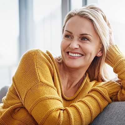 This is a color photograph featuring an adult woman with blonde hair leaning against a wall with her arm resting on it. She has a warm smile, wearing a yellow sweater, and appears relaxed.