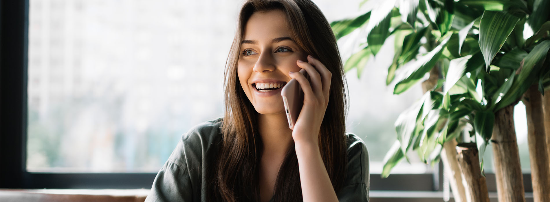 A young woman sitting at a table with a plant behind her, smiling and talking on her cell phone.
