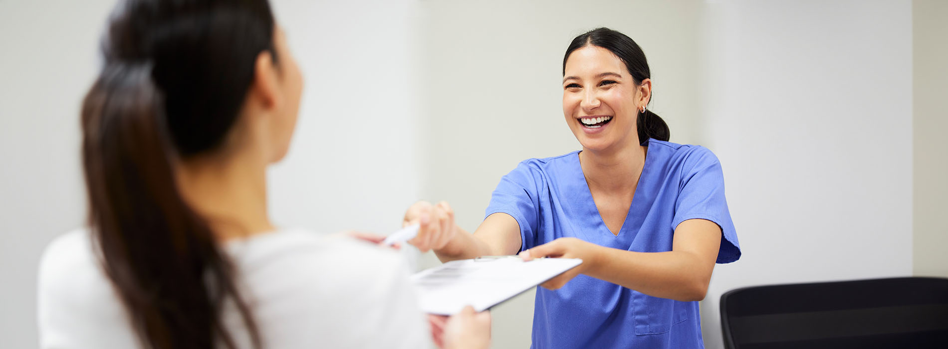 This image shows a woman in a white coat standing behind a desk with her hands on a clipboard, smiling and engaging with someone who appears to be handing her something  there s another person in the foreground looking at them both.
