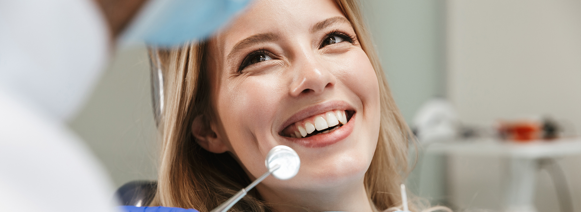 A woman with blonde hair smiling at a camera while seated in a dental chair, receiving dental care from a professional.