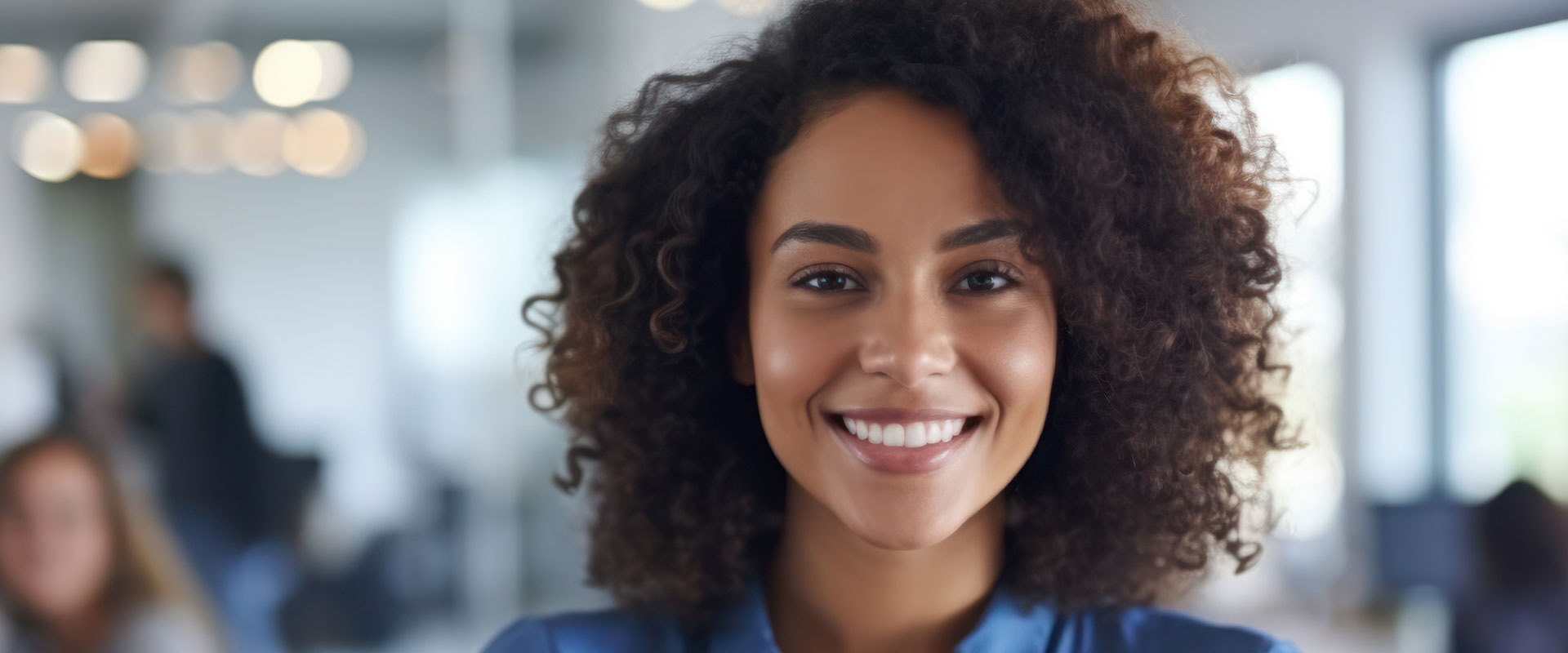 The image features a smiling individual with curly hair, wearing a dark shirt and glasses, against a blurred background that appears to be an office setting.