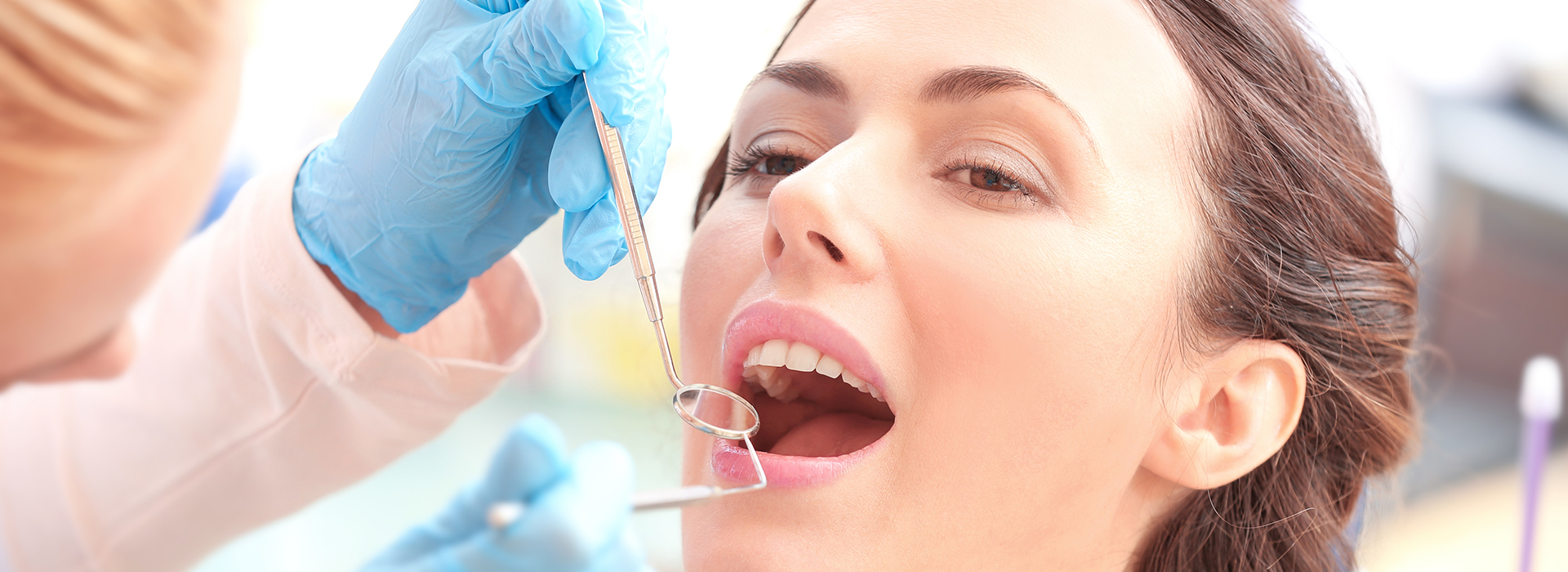 The image shows a woman receiving dental treatment under the care of a dentist, with a dental hygienist assisting and another person observing from behind. They are all wearing medical gloves and masks, emphasizing cleanliness and safety protocols.