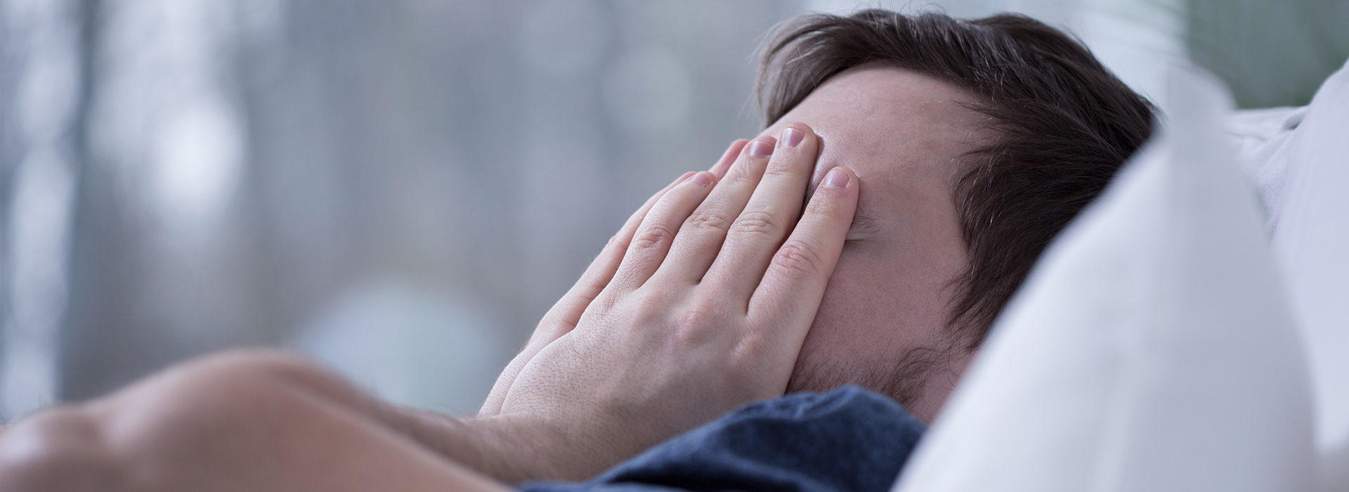 An adult male with his face covered by hands, lying on a bed.