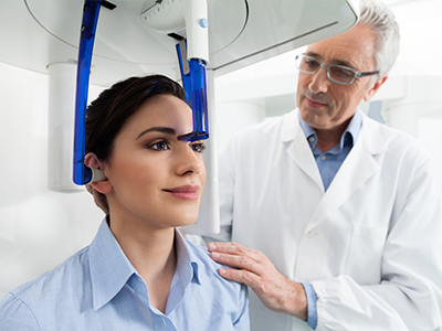 The image depicts a medical setting where a woman is seated with her head inside a device that appears to be a brain scanner, while a man in a white lab coat stands beside her, observing the process.