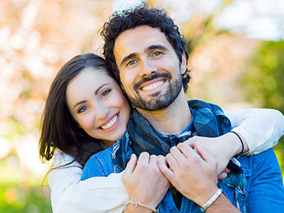A man and woman embracing each other outdoors, both smiling and looking at the camera.