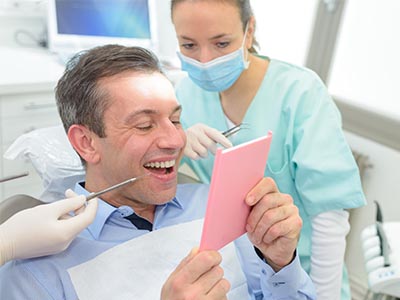 The image shows a man sitting in a dental chair with a surprised expression, holding a pink card with his name on it, while a dentist and dental hygienist look at him with smiles.