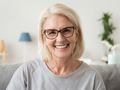 The image shows a woman with short blonde hair wearing glasses and a light-colored top, smiling at the camera while seated indoors.