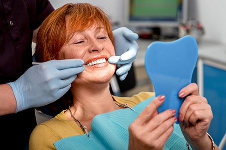 A woman sitting in a dental chair with her mouth open, receiving a teeth cleaning from a dental hygienist who is holding a mirror up to show her teeth.