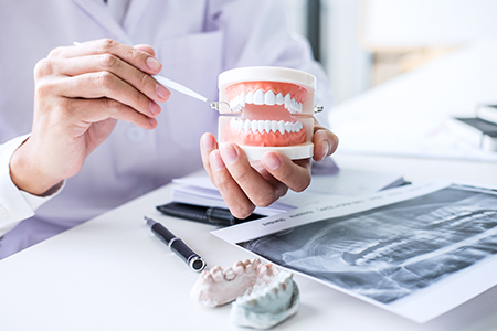The image shows two photographs side by side  on the left, a person s hand holding an open mouth model with a dental implant, and on the right, the same hand holding a cup of dental implants in front of a dental professional seated at a desk with various dental materials and equipment.