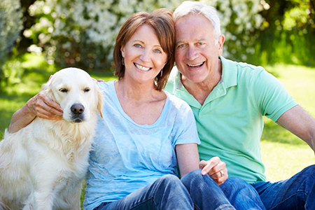 The image shows an older couple sitting outdoors with a golden retriever dog between them, smiling at the camera.