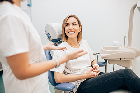 The image depicts a woman sitting in a dental chair, smiling at the camera, with a dental professional standing behind her, both in a dental office setting.