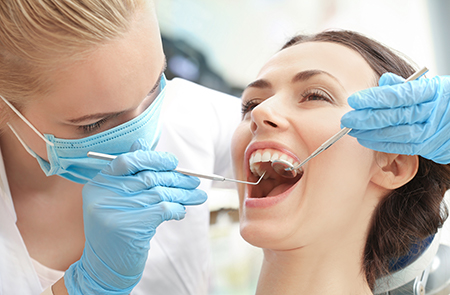 The image shows a dental hygienist performing a procedure on a patient s teeth while wearing protective gloves and a mask, with the patient sitting in a dental chair and holding their mouth open.
