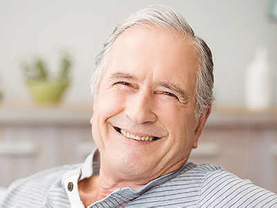 A smiling older man with gray hair, wearing a blue shirt, sitting comfortably in a cozy room.